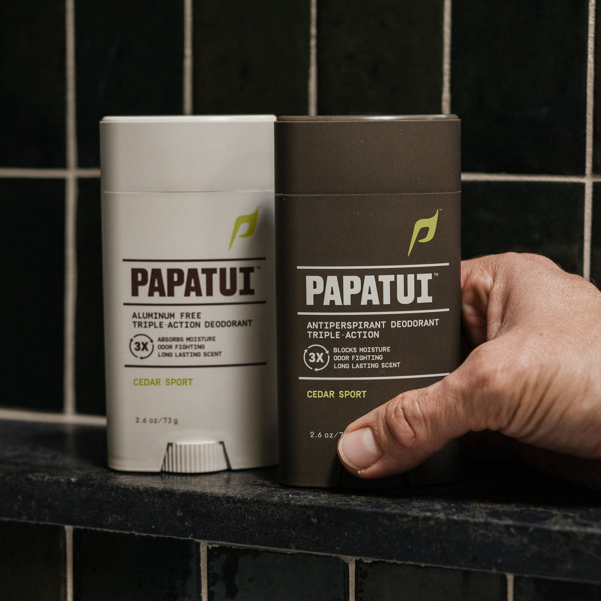 Hand holding PAPATUI Cedar Sport Antiperspirant Deodorant and Aluminum Free Deodorant against black tiles