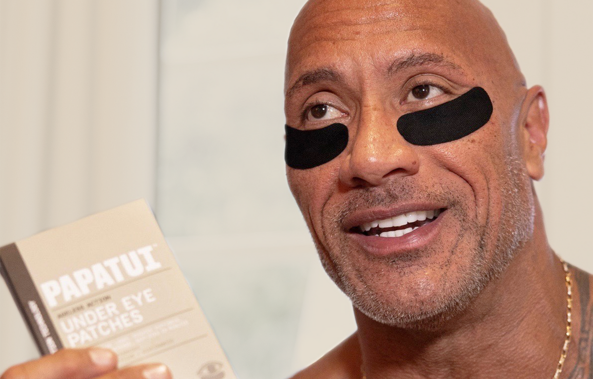 Dwayne "The Rock" Johnson wearing and holding a box of Papatui under eye patches against a neutral background