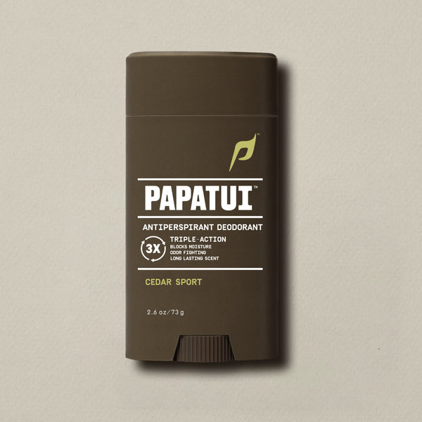 PAPATUI™ Antiperspirant Deodorant 2.6 oz in Cedar Sport scent. The product is housed in a dark brown stick with a minimalist beige and white label and light blue PAPATUI™ logo. Callouts on the label highlight triple-action benefits: blocks moisture, odor fighting, and long-lasting scent. Positioned against a neutral beige background.