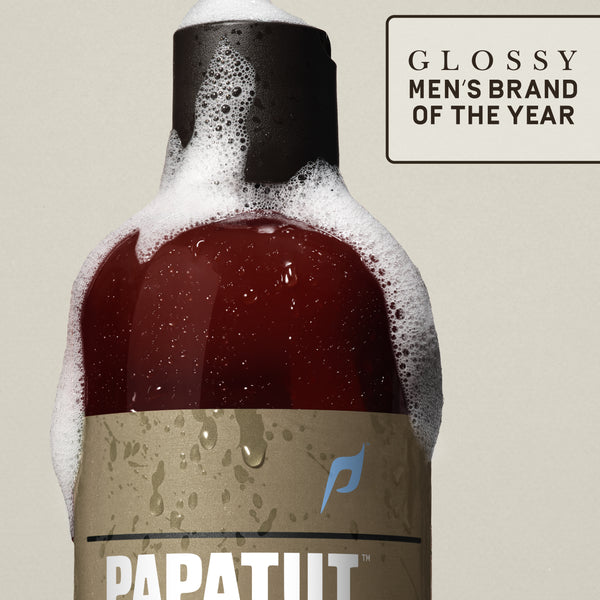 Close-up of PAPATUI™ Lush Coconut Refreshing Body Wash 18 fl oz bottle in a rich amber color with a black cap, covered in fresh cleansing foam. The bottle features a sleek minimalist beige label with the PAPATUI™ logo and is set against a soft tan background. The image includes a “Glossy Men’s Brand of the Year” badge.
