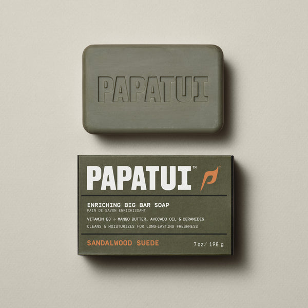 A bar Enriching Bar Soap in Cedar Sport scent rests on a beige background, with a sprig of greenery to the left. The soap is light beige and has a smooth texture with the PAPATUI brand name embossed on the surface.