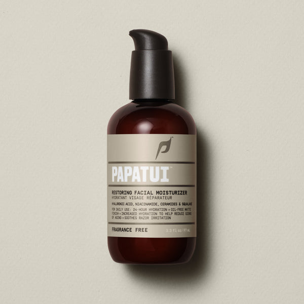 PAPATUI™ Unscented Restoring Facial Moisturizer 3.3oz front of pack set against a neutral beige background features a minimalist design with a brown-tinted bottle, black pump dispenser, and beige label with bold white text. The product contains hyaluronic acid, niacinamide, ceramides, and squalane, offering 24-hour hydration with an oil-free matte finish. It is fragrance-free and formulated to reduce signs of aging and soothe razor irritation.