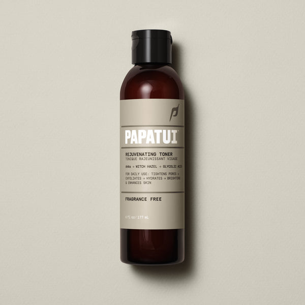 A sleek amber bottle of PAPATUI™ Unscented Rejuvenating Toner 6oz standing upright against a neutral beige background. The label features bold white branding with key product benefits, including hyaluronic acid, niacinamide, ceramides, and squalane for 24-hour hydration, an oil-free matte finish, and soothing razor irritation. The design highlights its fragrance-free formula, making it ideal for daily use and sensitive skin. The aesthetic is minimalist, modern, and premium front of pack.
