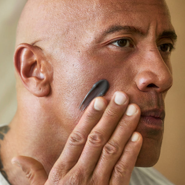 Dwayne Johnson applying PAPATUI™ Exfoliating Facial Scrub Unscented 4oz to his cheek. The dark gray scrub is spread smoothly on his skin as he gently rubs it in with his fingers. The close-up shot emphasizes the texture of the product and its exfoliating properties, with a neutral, softly lit background keeping the focus on his face and the scrub application.