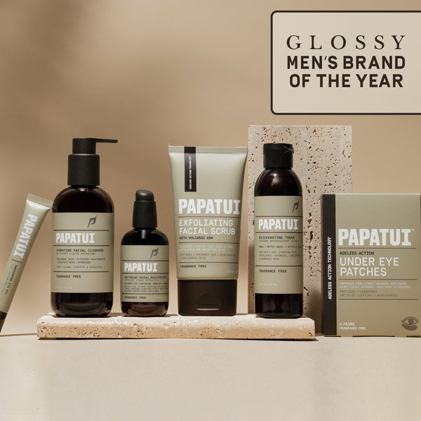 A display of PAPATUI™ mens skincare products, featuring a full lineup of the brand’s offerings. The set includes a face cleanser, exfoliating facial scrub, rejuvenating toner, under-eye patches, and eye gel, all in beige and black packaging with bold white branding. The background is a neutral-toned setting with textured stone elements, emphasizing a clean and premium aesthetic. A label in the top right corner highlights the brand’s recognition as ‘GLOSSY MEN’S BRAND OF THE YEAR.