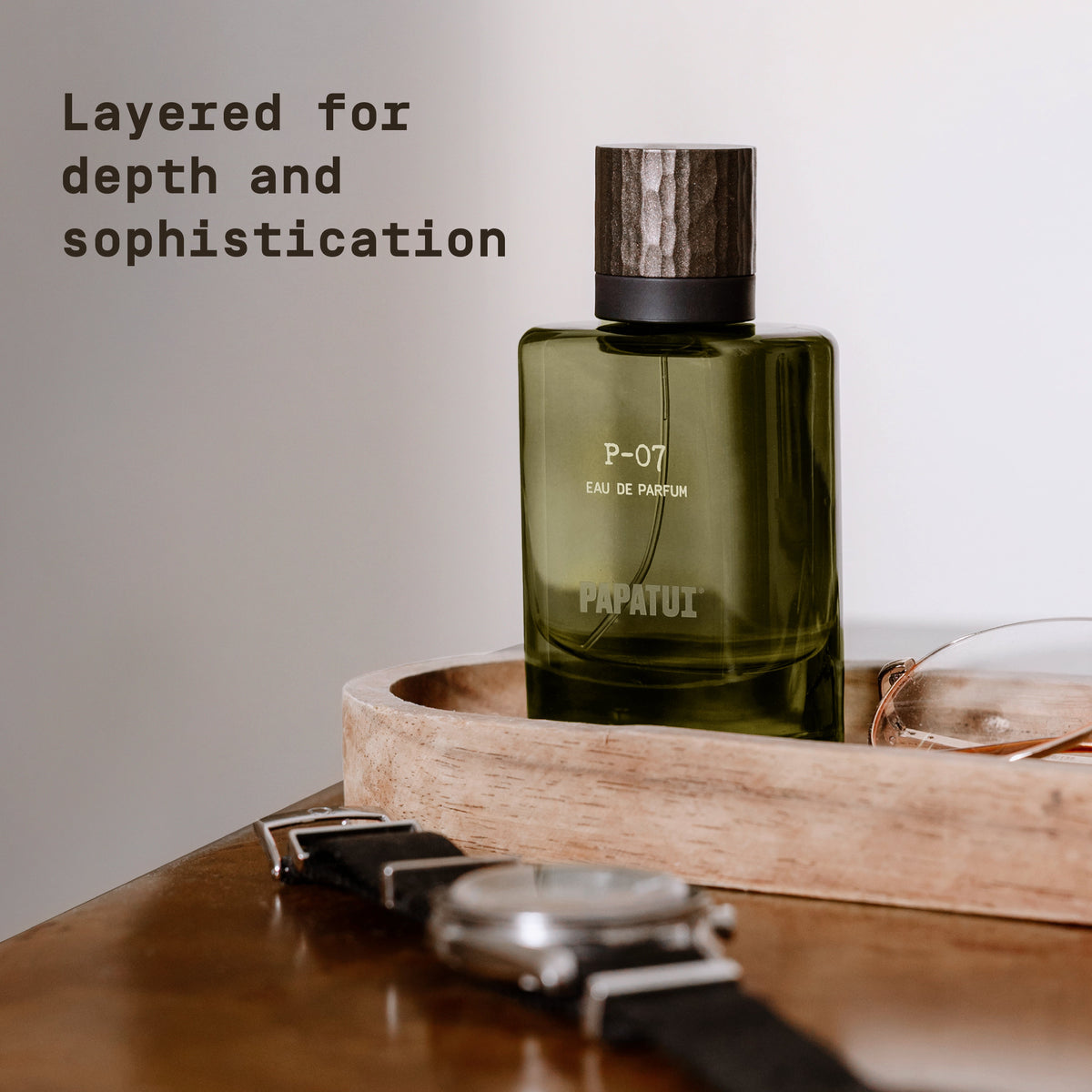 Green cologne bottle labeled 'P-07 Eau de Parfum' by 'PAPATUI' resting on a wooden surface next to a watch, with text overlay that reads "Layered for depth and sophistication"