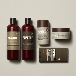 Flat lay of PAPATUI™ So Bright & So Clean Bundle in Vanilla Shea scent. Includes: Refreshing Body Wash, Nourishing Shampoo + Conditioner, Enhancing Tattoo Stick (Fragrance Free), Enhancing Tattoo Balm (Fragrance Free), and Antiperspirant Deodorant in Vanilla Shea. Products are arranged on a soft beige background with consistent amber and brown minimalist packaging.