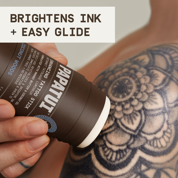 Hand applying PAPATUI™ Enhancing Tattoo Stick Coconut Woods 2.6 oz onto tattooed arm with floral black ink design. Showcases product's easy-glide formula to moisturize and enhance tattoo vibrancy.