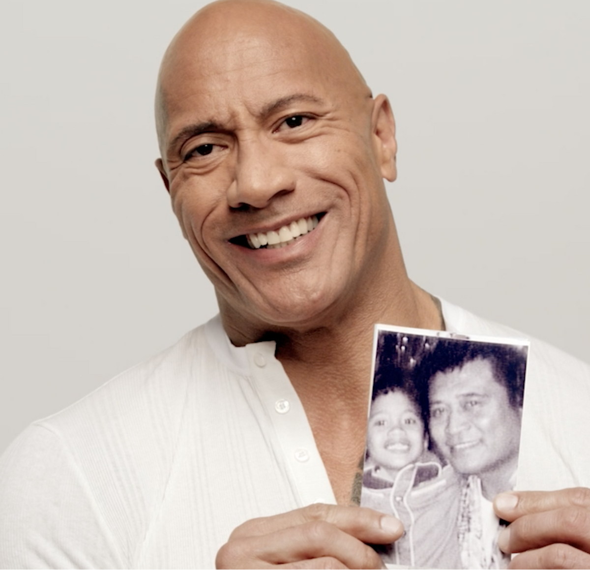 Dwayne "The Rock" Johnson holding a photo of himself as a child and his grandfather against a plain background