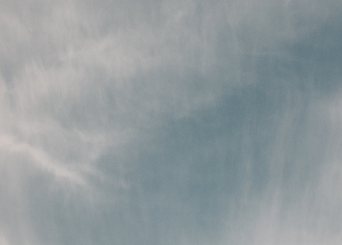 A soft-focus photograph of a cloudy sky with light gray and white clouds drifting across a pale blue background.