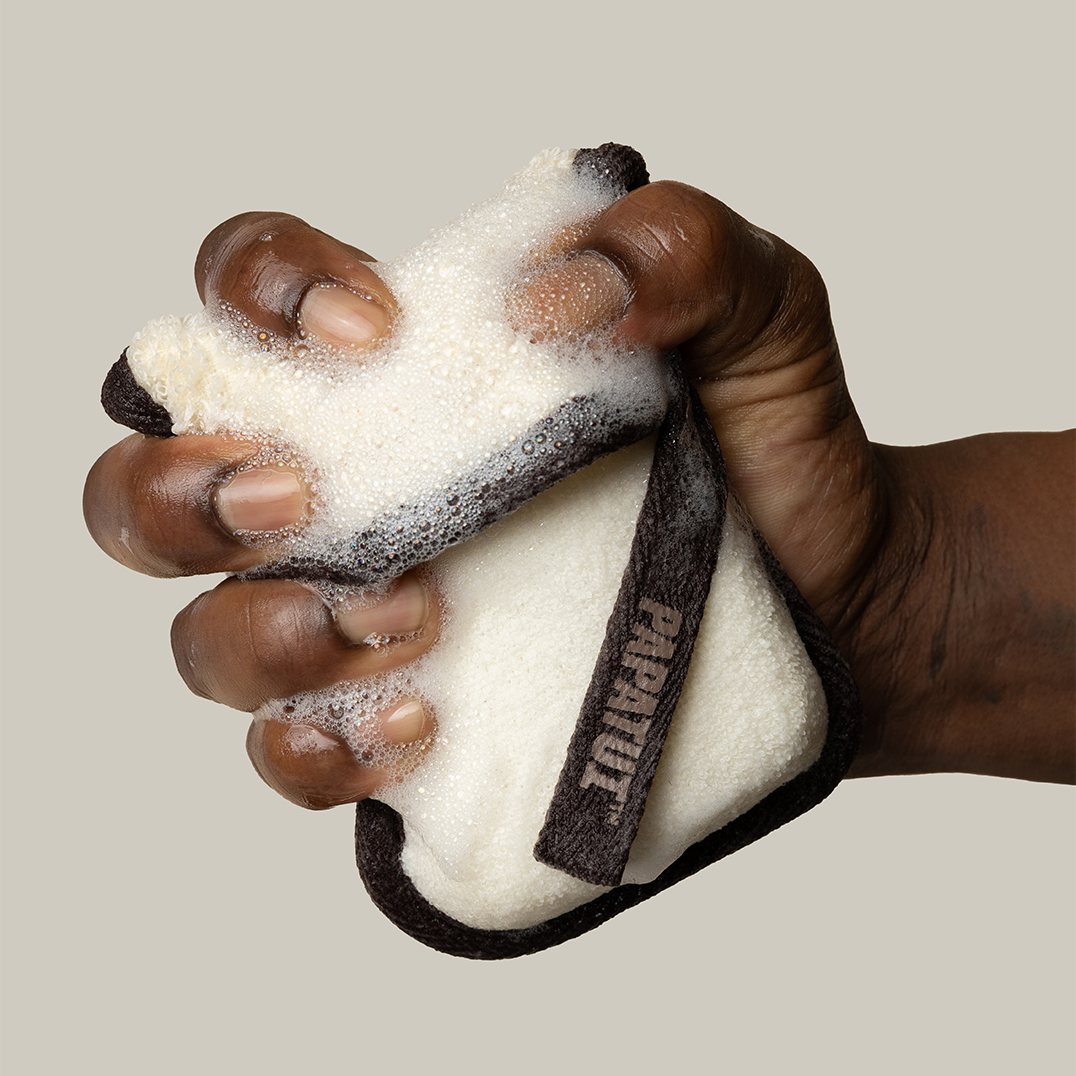 Hand holding sudsy PAPATUI Exfoliating Body Scrubber and Big Bar Soap against a neutral background