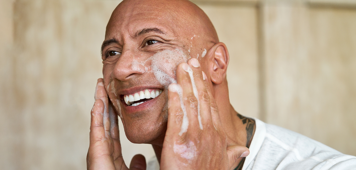 Dwayne "The Rock" Johnson applying soap to his face against a neutral background