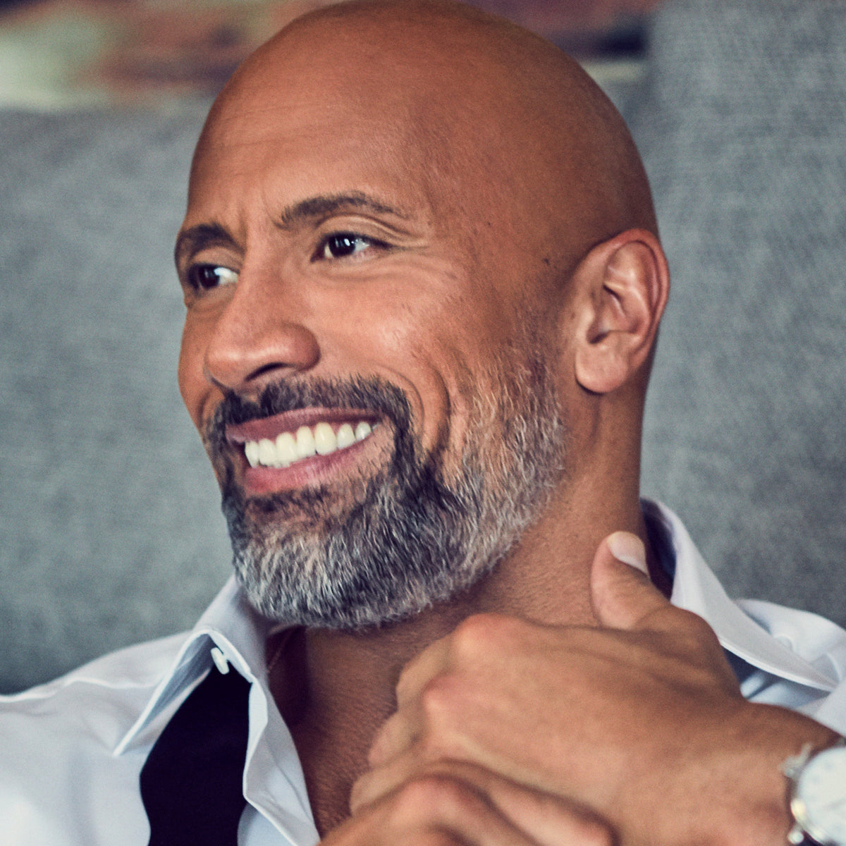 Dwayne "The Rock" Johnson sitting on a couch wearing a white shirt and black tie, smiling.