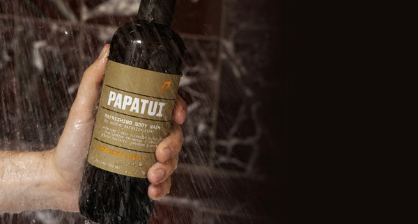 This image shows a hand holding Papatui 18 oz Sandalwood Suede Body Wash under running water in a shower with a marble background.