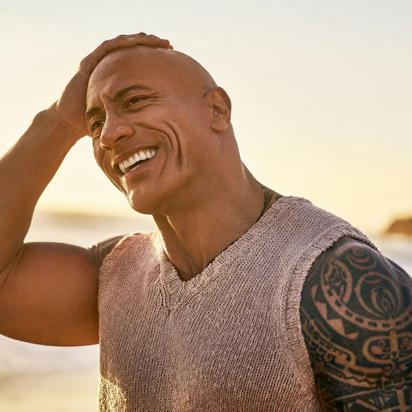 The image features Dwayne “The Rock” Johnson in a candid, joyful moment. He is seen laughing with a radiant smile, his right hand resting on his bald head. He is wearing a sleeveless, textured knit top in a neutral tone, revealing his muscular arms and intricate tribal tattoo on his left shoulder. The background is softly blurred with warm lighting, giving the image a natural, sunlit glow. His relaxed and charismatic expression exudes confidence and positivity. 