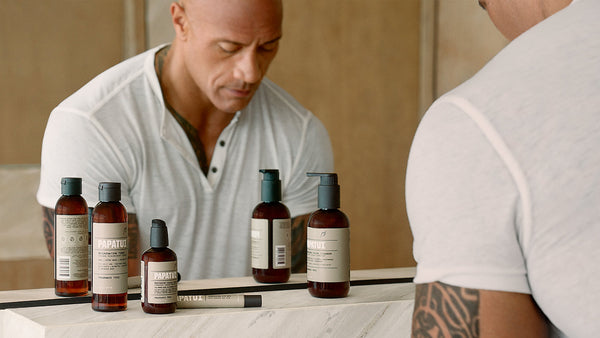 The image features Dwayne “The Rock” Johnson standing in front of a bathroom mirror, wearing a white short-sleeve shirt. On the bathroom counter in front of him is a lineup of PAPATUI™ mens skincare products.