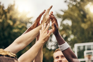 This image is a reflection of the PAPATUI™ Cares program which partners with youth group of diverse individuals. The image is engaging in a team huddle, raising their hands together for a high-five or unity gesture. The background is an outdoor setting with green trees and a blurred basketball hoop, suggesting a sports environment. hands are of various skin tones, symbolizing teamwork, inclusivity, and camaraderie. The warm lighting and natural backdrop create a motivational and uplifting setting.