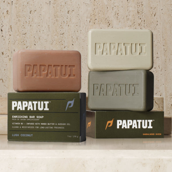 PAPATUI™ Enriching Big Bar Soap 7oz moisturizing mens bar soap enriched with Vitamin B3, mango butter, avocado oil, and ceramides. Designed to cleanse and hydrate for long-lasting freshness. The packaging is olive green with bold white typography, and the beige soap bar features an embossed "PAPATUI" logo front of pack featuring Lush Coconut, Sandalwood Suede, and Cedar Sport Fragrance.