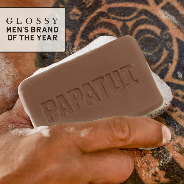 PAPATUI™ Lush Coconut Enriching Mens Big Bar Soap 7oz features a close-up of a hand holding the beige soap bar, which has the embossed "PAPATUI" logo, while lathering on tattooed skin. A "Glossy Men's Brand of the Year" badge is displayed in the upper left corner, emphasizing the brand’s recognition.