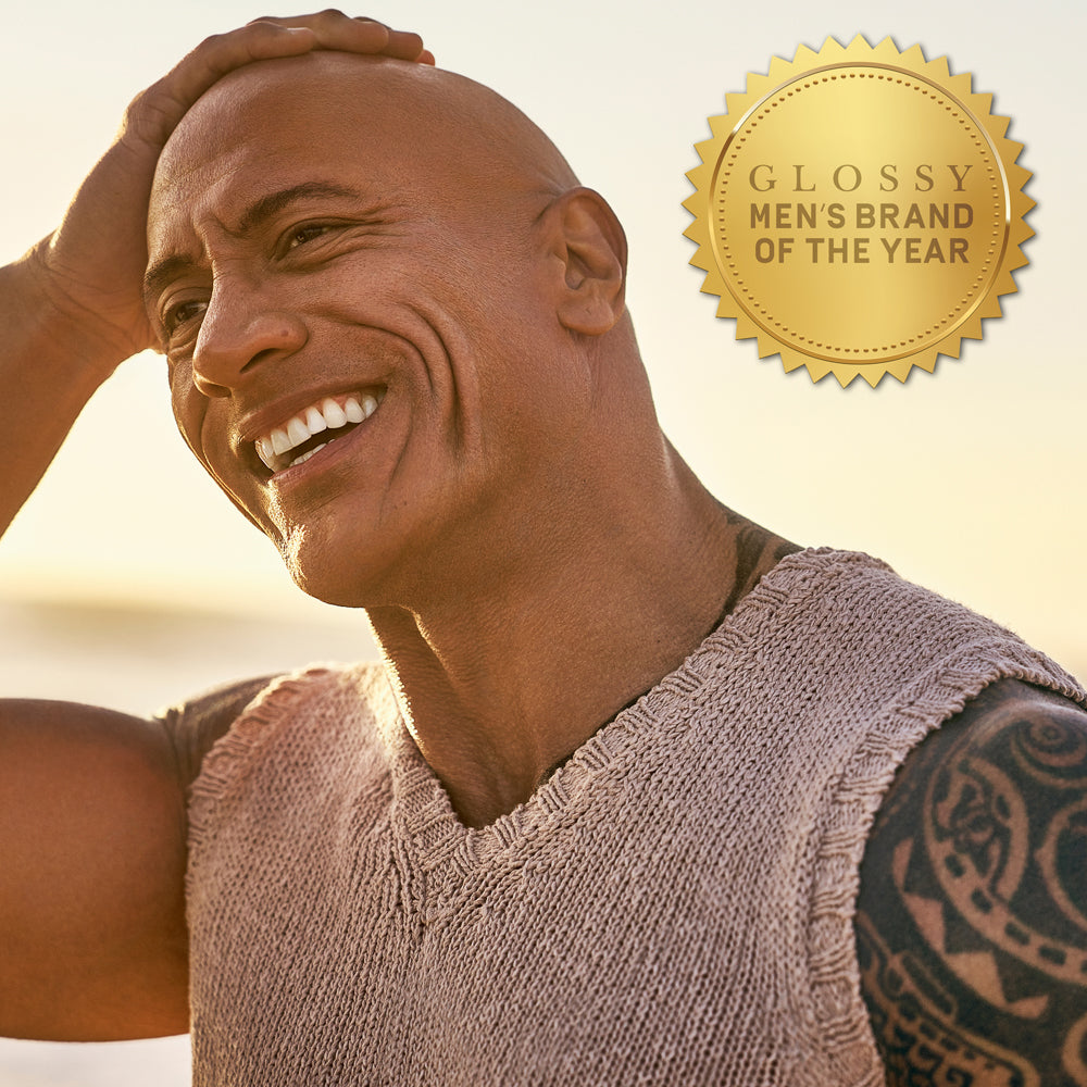Dwayne "The Rock" Johnson smiling outdoors, with gold award badge that reads "Glossy Men's Brand of the Year"