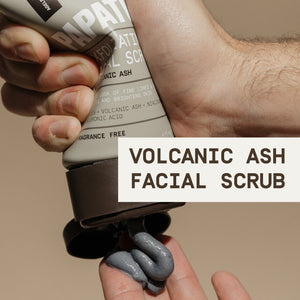 A close-up of a hand squeezing PAPATUI™ Exfoliating Facial Scrub onto another hand. The product is an unscented volcanic ash facial scrub in a 4oz tube, featuring a beige and brown design with bold white text. The thick, dark gray scrub is being dispensed, highlighting its exfoliating properties. The label mentions key ingredients such as volcanic ash, peptides, niacinamide, and hyaluronic acid, promoting skin brightening and minimizing fine lines and pores. A text overlay reads ‘Volcanic Ash Facial Scrub