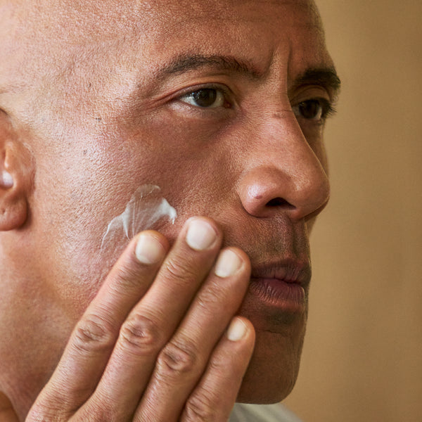 PAPATUI™ Unscented Hydrating Facial Cleanser 8 fl oz close-up shot of Dwayne Johnson applying PAPATUI™ Hydrating Facial Cleanser 8 fl oz to his cheek. The image highlights the smooth, hydrating texture of the cream as it absorbs into the skin. His skin appears healthy and moisturized, emphasizing the nourishing and anti-aging benefits of the product. The neutral, warm-toned background enhances the natural and effortless skincare routine.