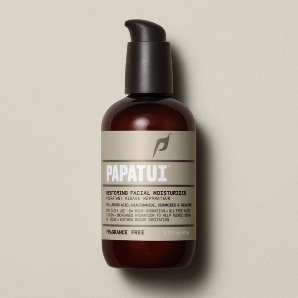 PAPATUI™ Unscented Restoring Facial Moisturizer 3.3oz front of pack set against a neutral beige background features a minimalist design with a brown-tinted bottle, black pump dispenser, and beige label with bold white text. The product contains hyaluronic acid, niacinamide, ceramides, and squalane, offering 24-hour hydration with an oil-free matte finish. It is fragrance-free and formulated to reduce signs of aging and soothe razor irritation.