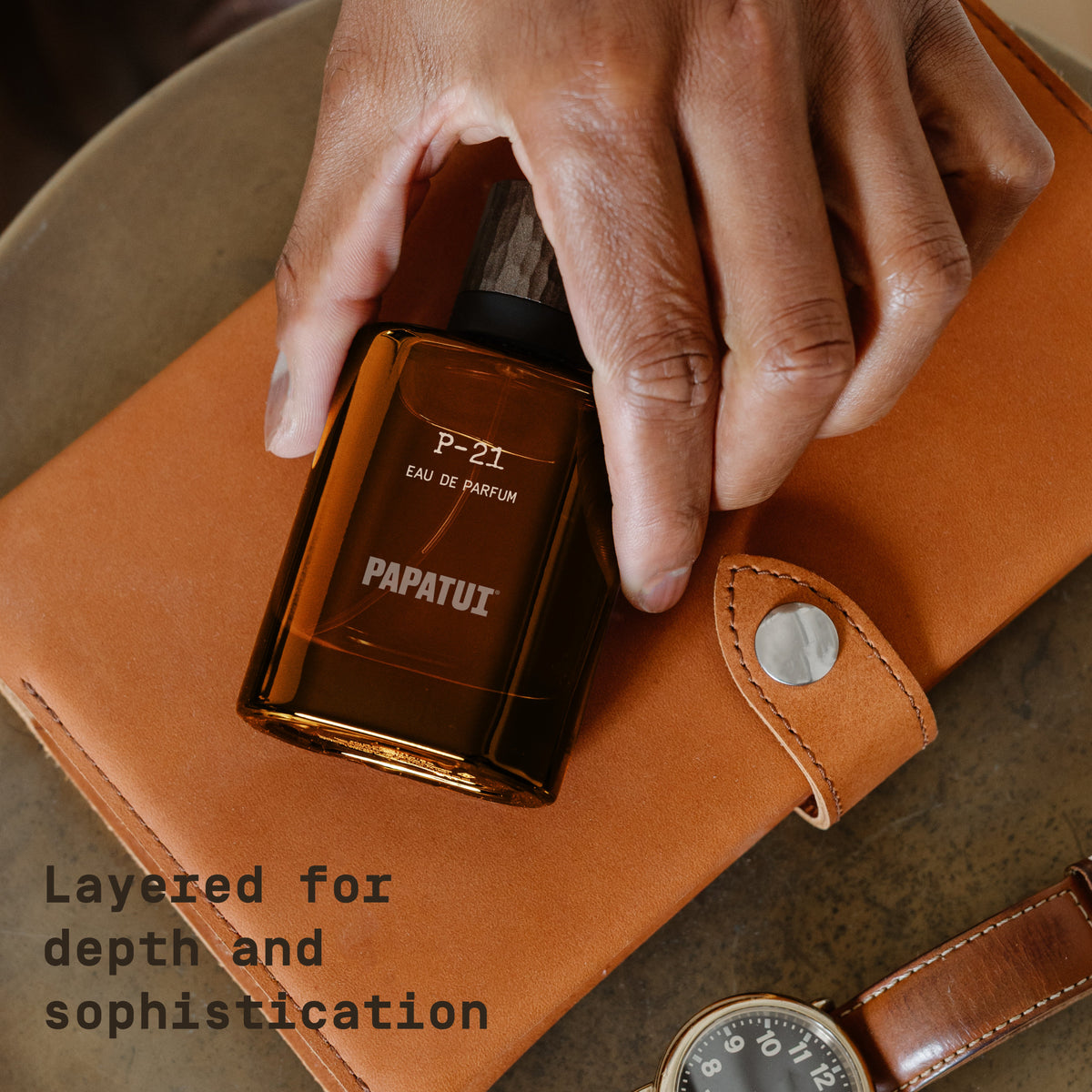 Hand holding amber cologne bottle labeled 'P-21 Eau de Parfum' by 'PAPATUI' on top of a brown leather case