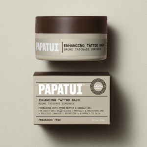 PAPATUI™ Enhancing Tattoo Balm in a 2 fl oz beige jar with a brown lid, placed above its matching packaging box. The label features bold white and black text, with the product name 'Enhancing Tattoo Balm' prominently displayed. The box provides additional details, highlighting key ingredients like mango butter and coconut oil, which help revitalize, protect, and brighten tattoo ink while delivering hydration and vibrancy to the skin. The formula is fragrance-free and designed for daily use.