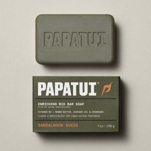 PAPATUI™ Sandalwood Suede Enriching Big Bar Soap 7oz moisturizing mens bar soap enriched with Vitamin B3, mango butter, avocado oil, and ceramides. Designed to cleanse and hydrate for long-lasting freshness. The packaging is olive green with bold white typography, and the beige soap bar features an embossed "PAPATUI" logo front of pack 