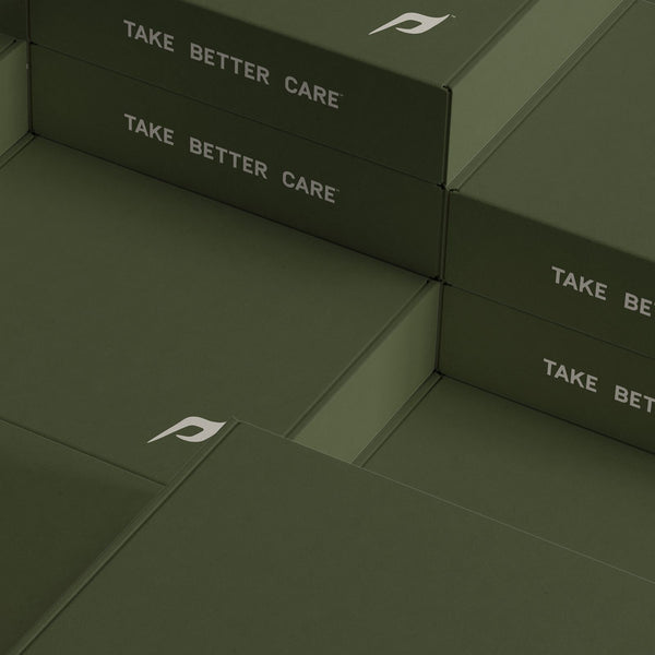 The image showcases a stack of olive-green boxes from Dwayne Johnson’s PAPATUI™ brand. Each box features the brand’s signature stylized “P” logo in white, placed on the upper corners. The phrase “TAKE BETTER CARE” is printed in bold, capitalized white letters across multiple boxes, reinforcing the brand’s self-care messaging. 