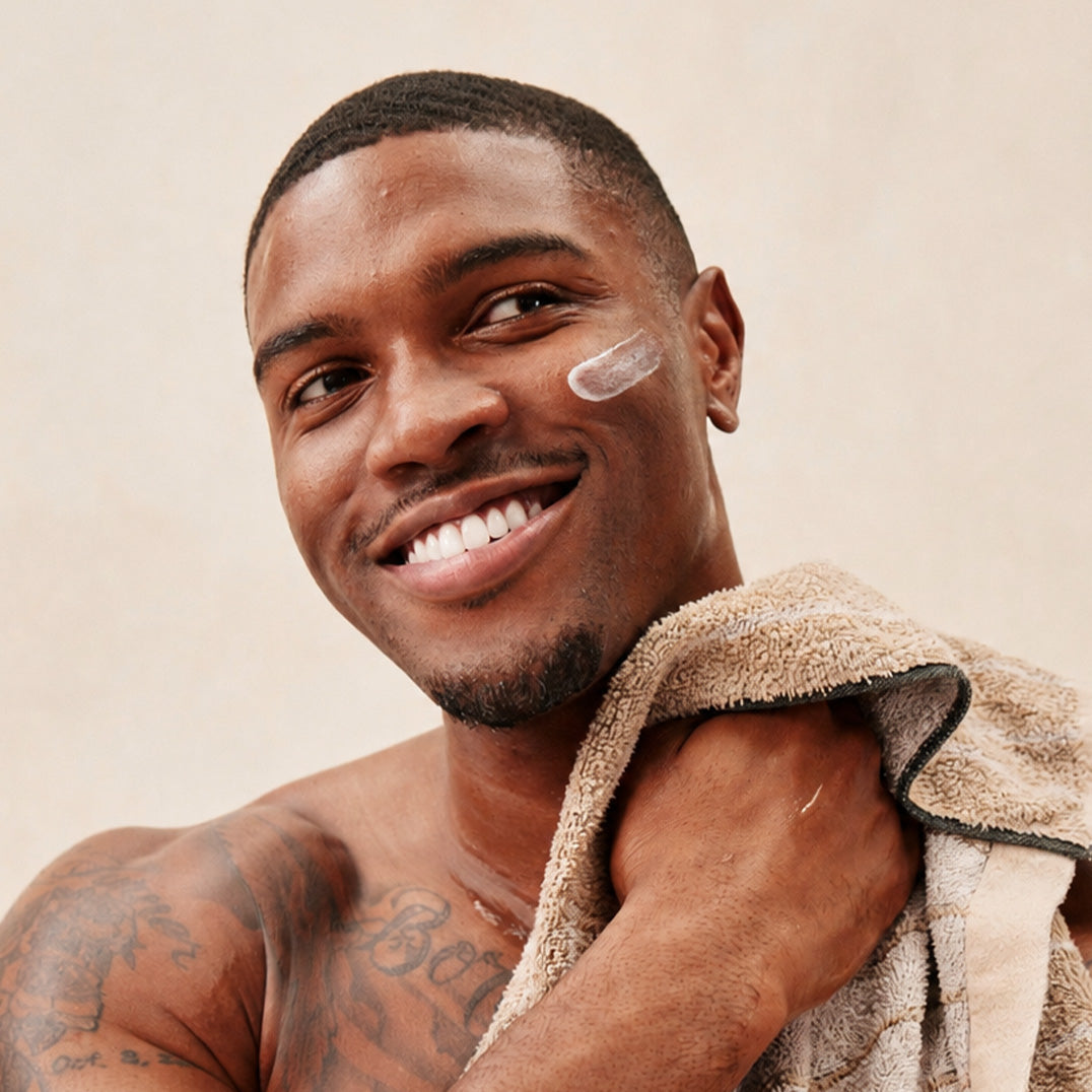 Man with a towel around his shoulder, smiling with a white cream on his cheek against a neutral background