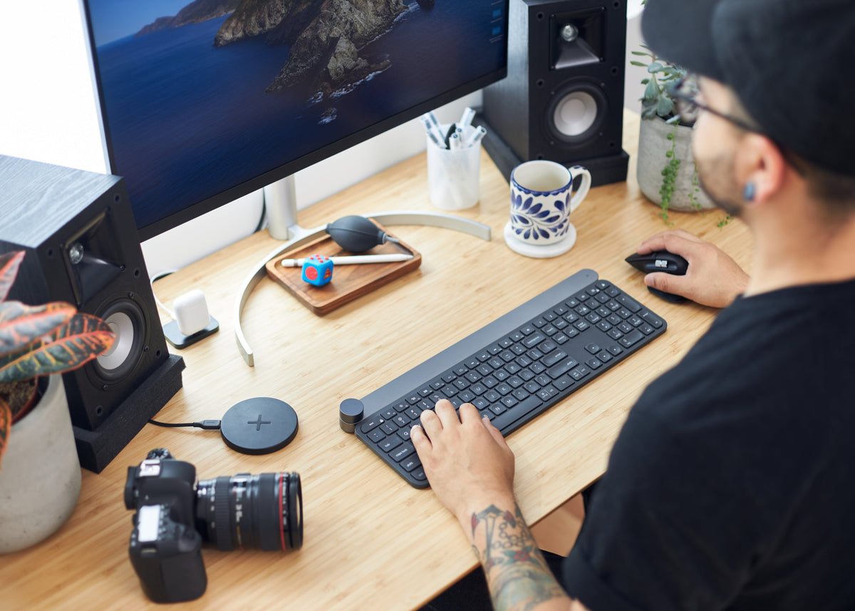 A man wearing a black cap and black t-shirt sits at a wooden desk, typing on a black wireless keyboard and using a mouse. The desk is styled with a computer monitor, speakers, a camera, a mug, and plants.