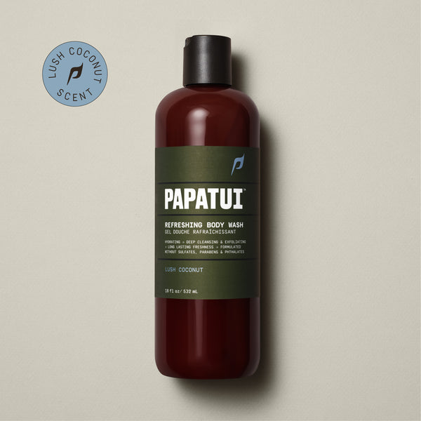 A PAPATUI Refreshing Body Wash in Lush Coconut scent in a translucent brown bottle with a black cap, displayed against a beige background with a few dark twigs. The label on the bottle is in a matching brown tone with white and orange text, highlighting features like hydrating, deep cleansing, exfoliating, and long-lasting freshness.