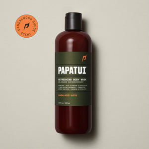 A PAPATUI Refreshing Body Wash in Sandalwood Suede scent in a translucent brown bottle with a black cap, displayed against a beige background with a few dark twigs. The label on the bottle is in a matching brown tone with white and orange text, highlighting features like hydrating, deep cleansing, exfoliating, and long-lasting freshness.