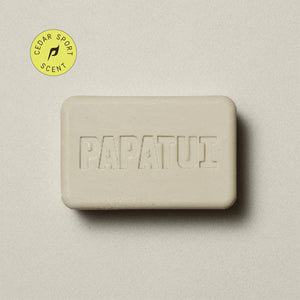 A bar Enriching Bar Soap in Cedar Sport scent rests on a beige background, with a sprig of greenery to the left. The soap is light beige and has a smooth texture with the PAPATUI brand name embossed on the surface. 