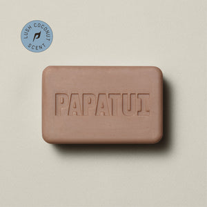 A bar Enriching Bar Soap in Lush Coconut scent rests on a beige background, with a sprig of greenery to the left. The soap is light beige and has a smooth texture with the PAPATUI brand name embossed on the surface. 