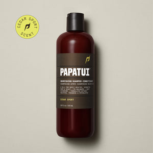 A bottle of PAPATUI Nourishing Shampoo-Conditioner in Cedar Sport scent is showcased on a beige background, accompanied by a sprig of greenery to the left. The bottle is translucent brown with a black cap and a gray-black label that includes the PAPATUI logo in white and yellow. The volume stated is 18 fl oz or 532 ml.