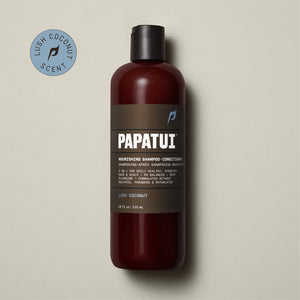 A bottle of PAPATUI Nourishing Shampoo-Conditioner in Lush Coconut scent is showcased on a beige background, accompanied by a sprig of greenery to the left. The bottle is translucent brown with a black cap and a gray-black label that includes the PAPATUI logo in white and yellow. The volume stated is 18 fl oz or 532 ml.