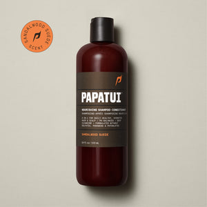 A bottle of PAPATUI Nourishing Shampoo-Conditioner in Sandalwood Suede scent is showcased on a beige background, accompanied by a sprig of greenery to the left. The bottle is translucent brown with a black cap and a gray-black label that includes the PAPATUI logo in white and yellow. The volume stated is 18 fl oz or 532 ml.