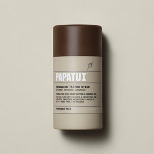 A PAPATUI Enhancing Tattoo Stick against a beige background. The product comes in a compact, brown twist-up container with a lighter tan label. It's formulated with mango butter and coconut oil, designed to revitalize and brighten tattoo ink. The label notes that it's fragrance-free, provides immediate hydration and sheen to tattoos, and leaves no residue. The size of the stick is 2.6 oz or 73 g.