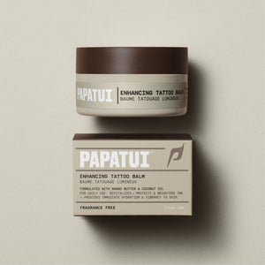 A jar of PAPATUI Enhancing Tattoo Balm positioned above its packaging box on a beige background. The jar has a brown lid and a pale green label with the PAPATUI logo in white. It is formulated with mango butter and coconut oil for daily use, to revitalize, protect, and brighten tattoos while providing immediate hydration and vibrancy to the skin. The product is fragrance-free and the jar contains 2 fl oz or 59 ml of balm.