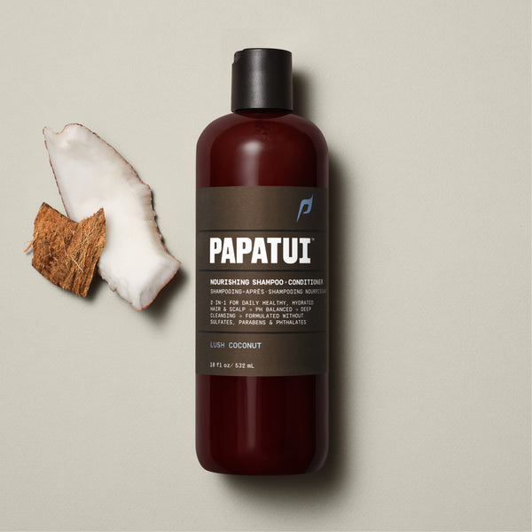 A bottle of PAPATUI Nourishing Shampoo-Conditioner in Lush Coconut scent is showcased on a beige background, accompanied by a sprig of greenery to the left. The bottle is translucent brown with a black cap and a gray-black label that includes the PAPATUI logo in white and yellow. The volume stated is 18 fl oz or 532 ml.