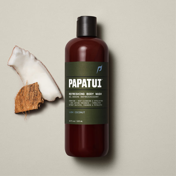 A PAPATUI Refreshing Body Wash in Lush Coconut scent in a translucent brown bottle with a black cap, displayed against a beige background with a few dark twigs. The label on the bottle is in a matching brown tone with white and orange text, highlighting features like hydrating, deep cleansing, exfoliating, and long-lasting freshness.