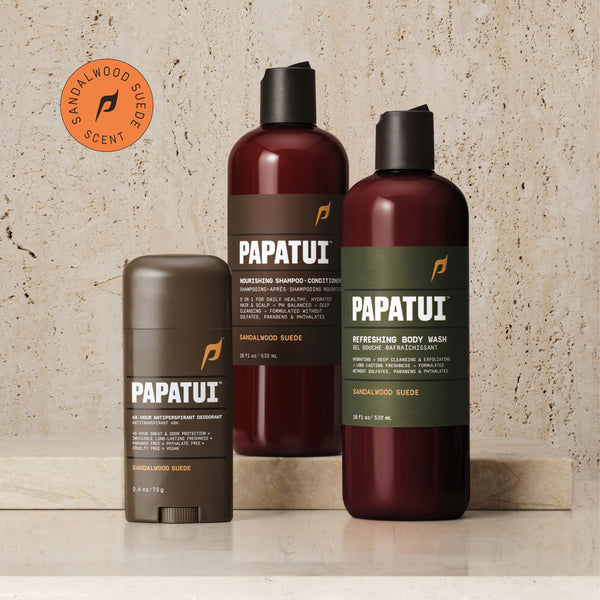 A PAPATUI men's personal care bundle displayed on a marble surface. The set includes a 48-Hour Antiperspirant Deodorant in Sandalwood Suede scent in a brown stick form, a Nourishing Shampoo-Conditioner, and a Refreshing Body Wash, both in Sandalwood Suede scent, housed in translucent brown bottles with black caps and green labels detailing their benefits.