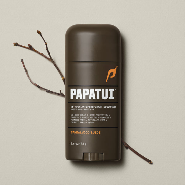 A dark brown stick of PAPATUI 48-Hour Antiperspirant Deodorant in Sandalwood Suede scent, positioned on a beige background with small dark twigs to the side. The product features orange and white text on the label, promising 48-hour sweat and odor protection, invisibility on clothes, and long-lasting freshness. The deodorant's size is indicated as 2.6 ounces or 73 grams.