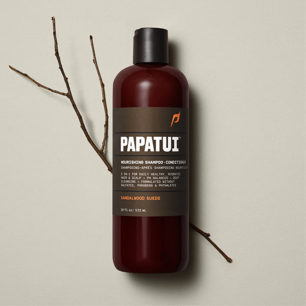 A bottle of PAPATUI Nourishing Shampoo-Conditioner in Sandalwood Suede scent is showcased on a beige background, accompanied by a sprig of greenery to the left. The bottle is translucent brown with a black cap and a gray-black label that includes the PAPATUI logo in white and yellow. The volume stated is 18 fl oz or 532 ml
