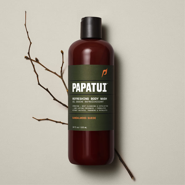 A PAPATUI Refreshing Body Wash in Sandalwood Suede scent in a translucent brown bottle with a black cap, displayed against a beige background with a few dark twigs. The label on the bottle is in a matching brown tone with white and orange text, highlighting features like hydrating, deep cleansing, exfoliating, and long-lasting freshness.