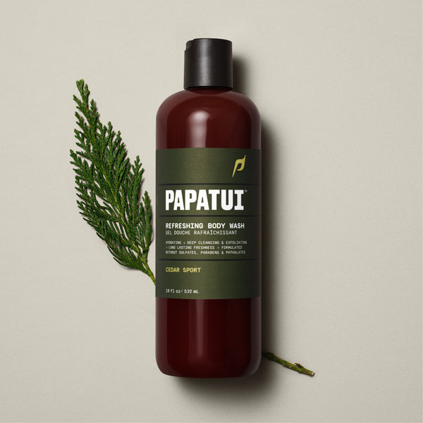 A PAPATUI Refreshing Body Wash in Cedar Sport scent in a translucent brown bottle with a black cap, displayed against a beige background with a few dark twigs. The label on the bottle is in a matching brown tone with white and orange text, highlighting features like hydrating, deep cleansing, exfoliating, and long-lasting freshness.